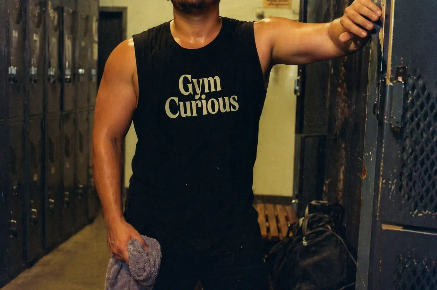 Jock in locker room wearing ‘Gym Curious’ black muscle tank